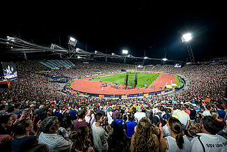 Stade olympique de Munich plein à l’Euro d’athlétisme 2022. Piste rouge éclairée et terrain central de nuit. Stade olympique de Munich lors des Championnats d’Europe d’athlétisme 2022 en soirée. La piste rouge en revêtement synthétique est éclairée. Des milliers de spectateurs assistent aux compétitions d’athlétisme sous les projecteurs.
