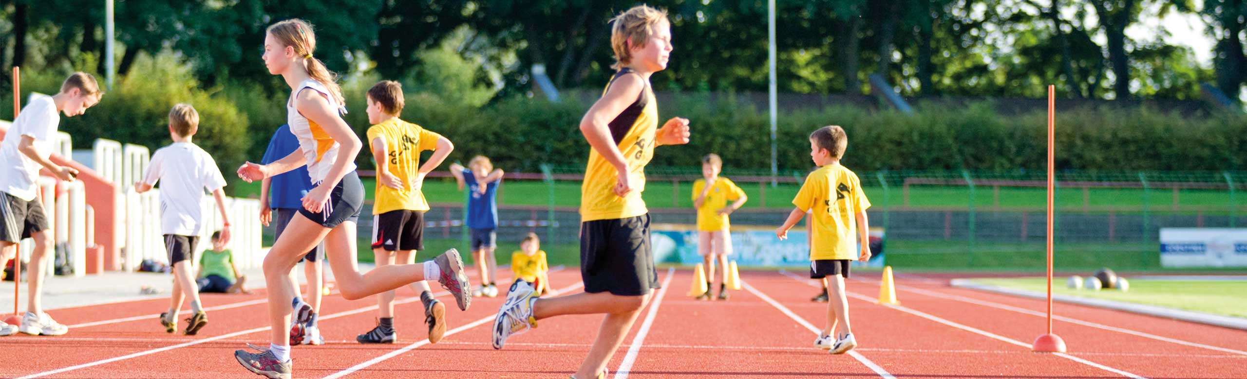 Children and young athletes running and playing on a red synthetic track under sunny skies at the stadium.