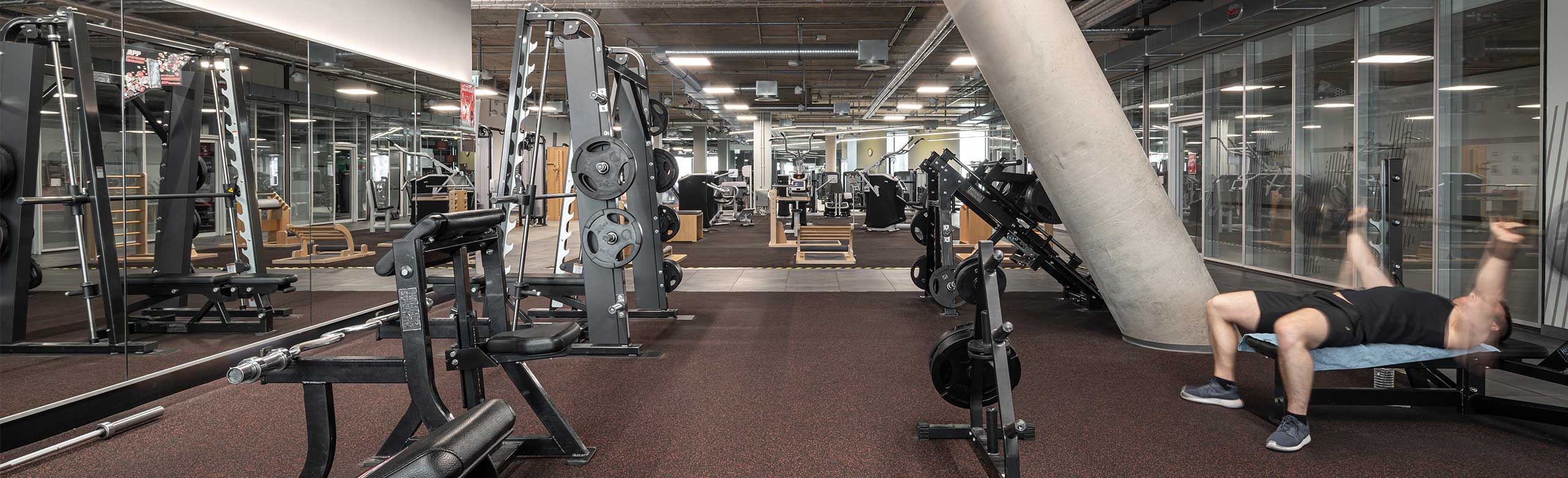 Revêtement de sol REGUPOL dans une zone d'entraînement aux haltères libres dans un centre de fitness. On y voit un homme allongé sur un banc de musculation en train de soulever des haltères.