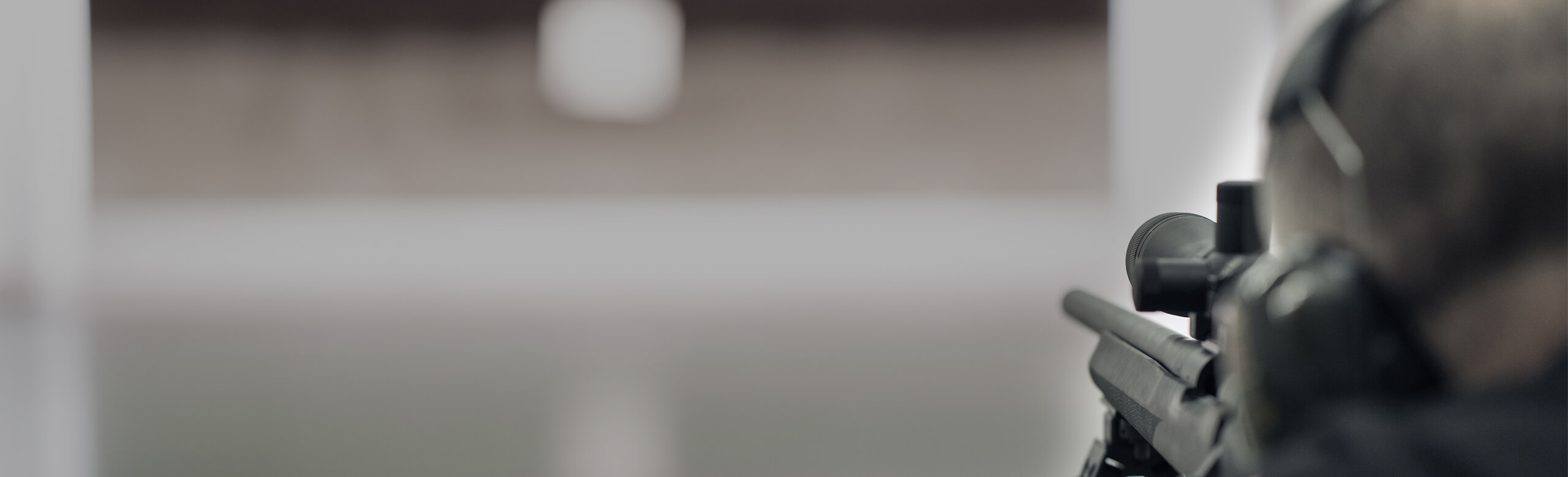 A shooter with headphones aims a rifle at a distant target in a shooting range.