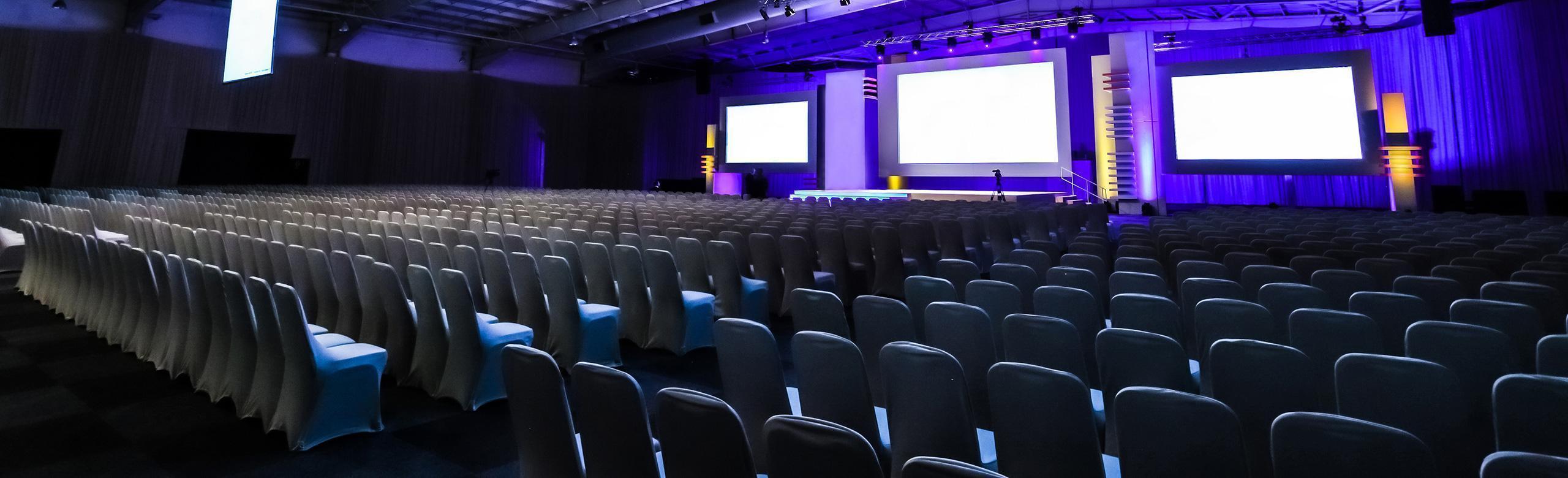 De nombreuses rangées de chaises orientées vers la scène. De nombreuses rangées de chaises orientées vers la scène. La scène se trouve dans un gymnase et est rétroéclairée par une lumière violette. Trois grands écrans sont accrochés au mur de la scène.