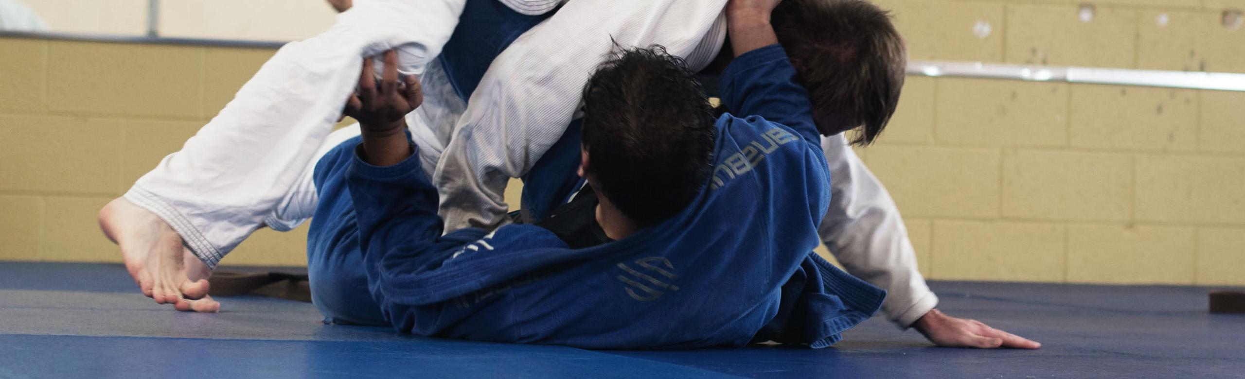 Deux personnes portent des combinaisons de jiu-jitsu et se trouvent en combat au sol sur des tapis bleus dans une salle de sport.