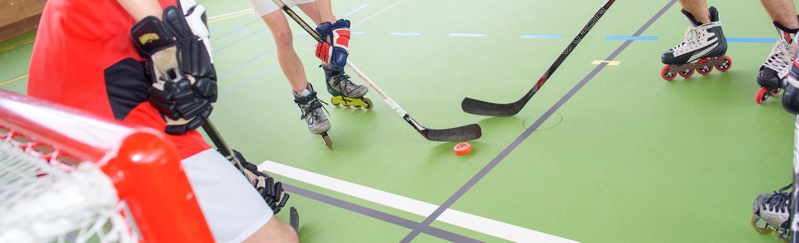 Différentes personnes jouent au hockey en ligne dans une salle de sport sur un sol vert de salle de sport avec différents lignages. Ils portent des patins en ligne et des gants de hockey, ont des crosses de hockey dans les mains et se passent le palet devant un but. 