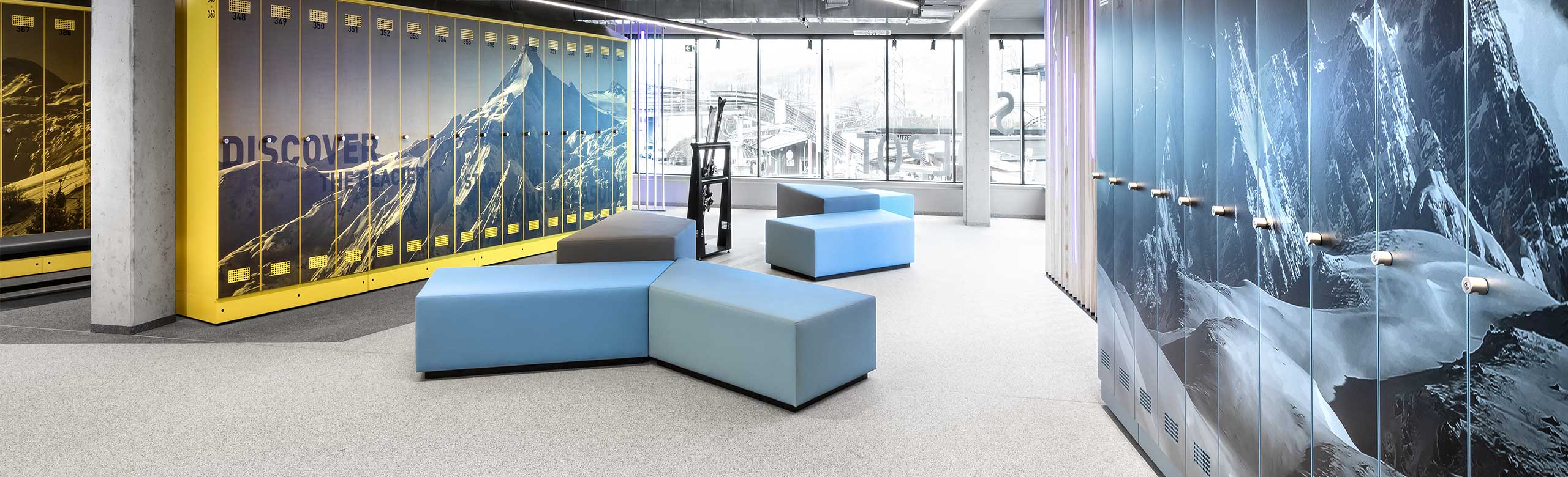Un dépôt de skis lumineux dans une station de remontées mécaniques. Au fond de la pièce se trouve une grande baie vitrée, à droite et à gauche de l'image, on voit des casiers avec des motifs de montagne. Au milieu se trouvent plusieurs sièges de couleur bleue et grise. Le revêtement de sol est clair, en dessous des casiers, il est gris foncé.