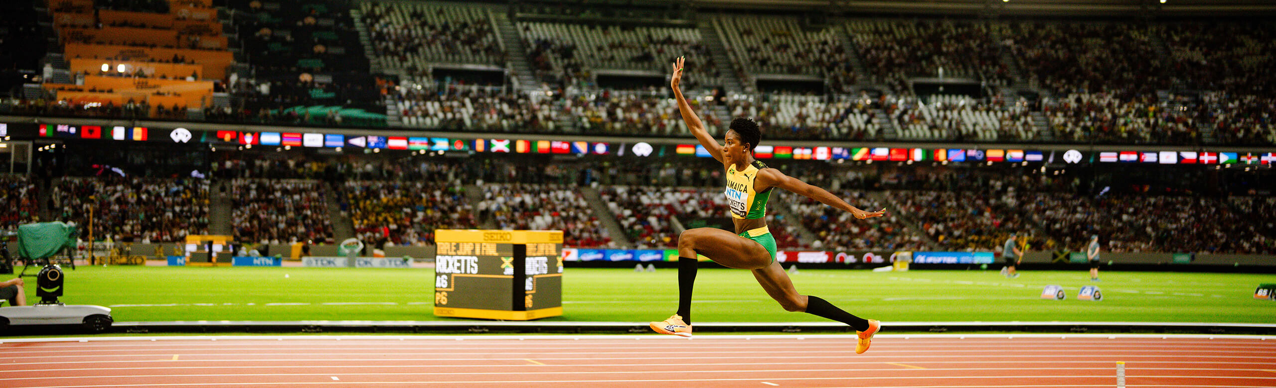 Triple saut d’une athlète lors d’un concours international d’athlétisme sur piste synthétique éclairée. Athlète féminine en plein saut lors d’un concours international de triple saut dans un stade rempli. La piste d’élan est en revêtement synthétique coloré, sous les projecteurs du stade.