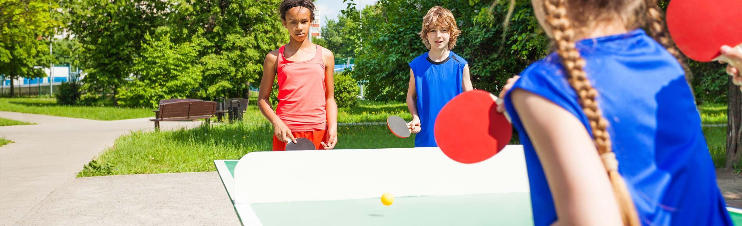 Children play table tennis on a playground with large format REGUPOL playfix elastic tiles, surrounded by trees - for safe playgrounds