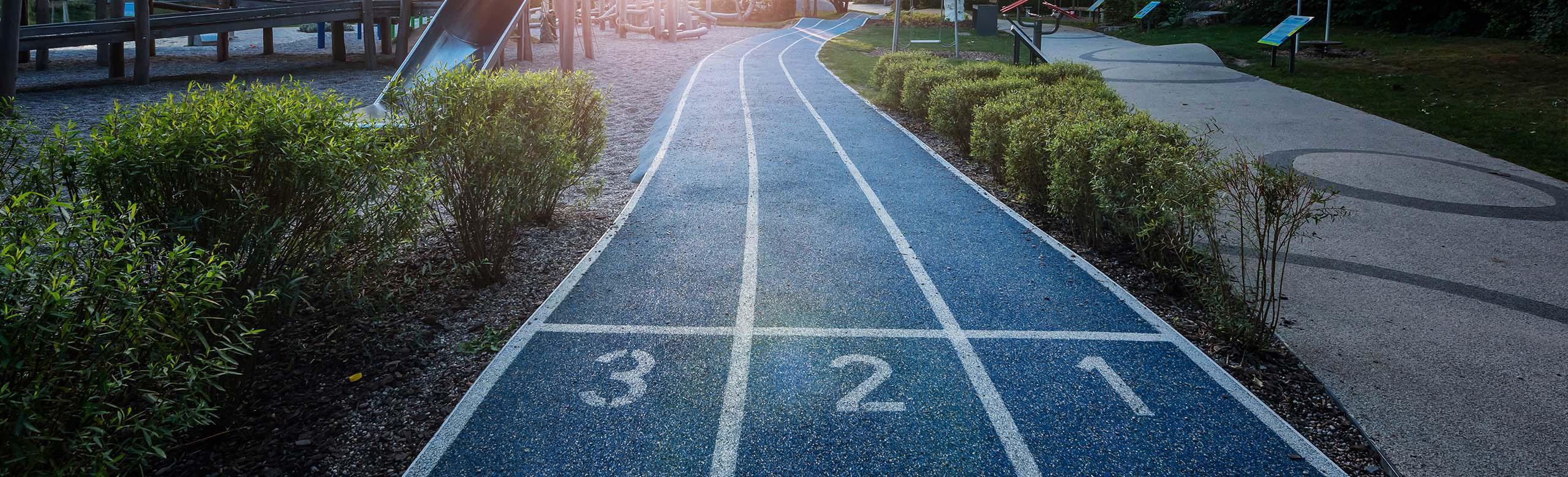 Playground with blue seamless REGUPOL safety flooring, marked with the numbers 3, 2, 1 and resembling a running track. The track winds its way through a green park landscape with bushes and play equipment in the background.