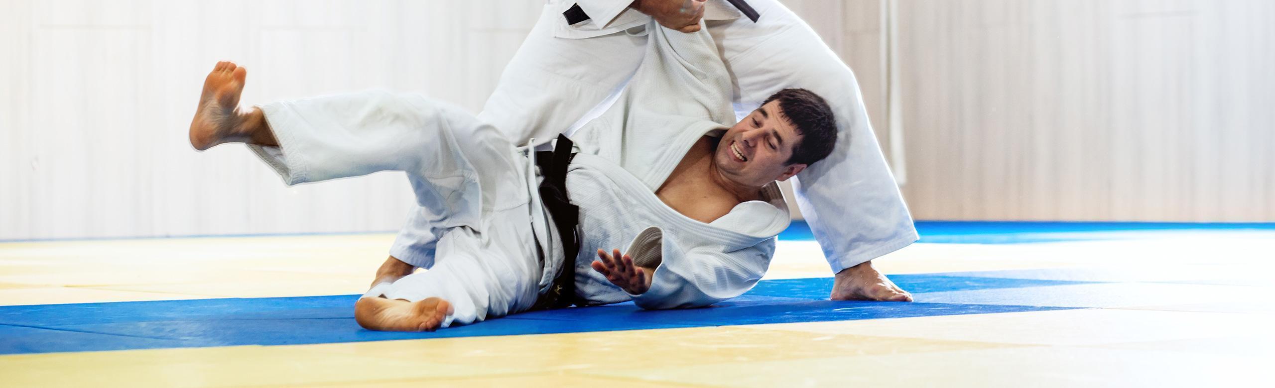 Two judokas fight on a blue and yellow tatami mat; one is being thrown to the ground.